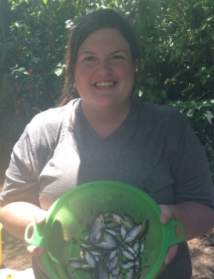 Grace is looking into the camera and smiling. There are tropical trees in the background and she is wearing a gray tshirt and holding a green sive full of Rhoadsia altipinna, a small western Ecuadorian Tetra fish which appear rainbow.